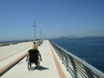 Donna con disabilità in carrozzina, fotografata di spalle in un percorso in riva al mare