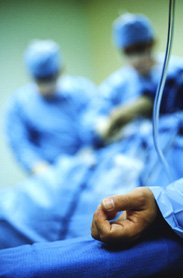 In primo piano braccio con flebo di persona malata su un letto di ospedale; sullo sfondo medici sfuocati