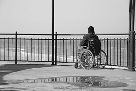 Foto in bianco e nero di persona in carrozzina, fotografata di spalle, mentre guarda il mare