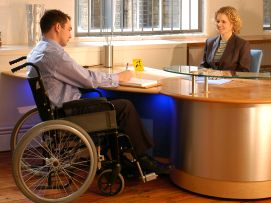 Uomo con disabilità davanti a una scrivania. Dietro un'operatrice non disabile