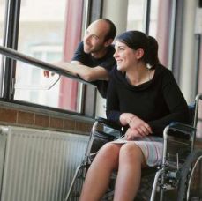 Giovane donna in carrozzina alla finestra, insieme a un giovane uomo non disabile