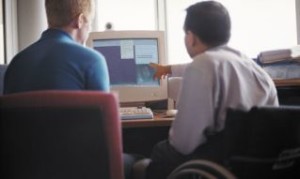 Giovane in carrozzina fotografato di spalle, lavora al computer insieme a uomo non disabile