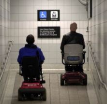 Due persone in carrozzina, di spalle, nel corridoio di una struttura sanitaria