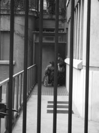 In primo piano sbarre di un cancello, sullo sfondo una persona in carrozzina con l'accompagnatore. Immagine in bianco e nero