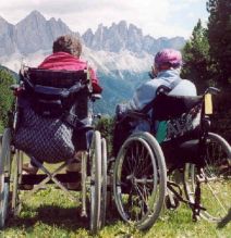 Due persone in carrozzina in montagna, fotografate di spalle